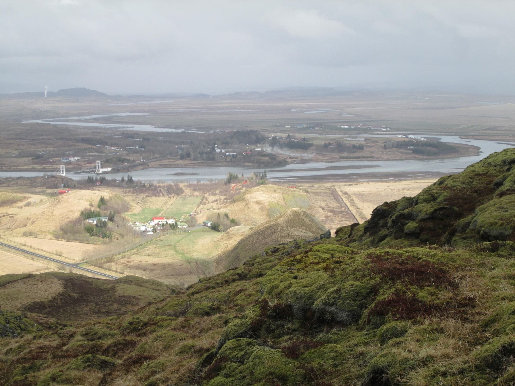 overview of a landscape and area of Laugarás, Bláskógarbyggð. Hvítá river seen and bridge going over, some lava and grass, trees and some houses